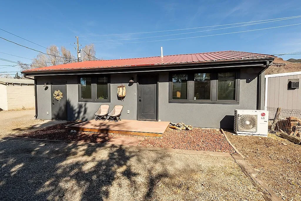 Backyard of a gray house with black window frames and a red metal roof, featuring a wooden patio with two chairs, an air conditioning unit, and leafless trees in the background.
