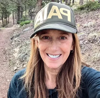 woman smiling outdoors in front of trees, wearing a black Patagonia cap and a black shirt