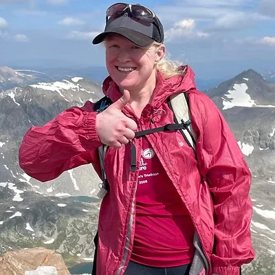 A woman in a red jacket giving a thumbs-up, standing on a mountainous landscape with snow patches and cloudy sky behind her.
