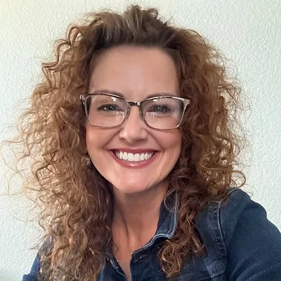 A woman with curly hair and glasses smiling, wearing a denim jacket against a plain wall.