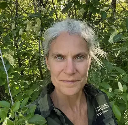 A woman with gray hair taking a selfie outdoors among green leaves and foliage.