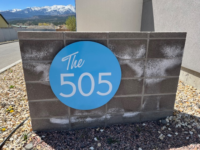 A stone sign with a blue circle displaying the words 'The 505' in white text, situated outdoors with a mountainous landscape in the background.