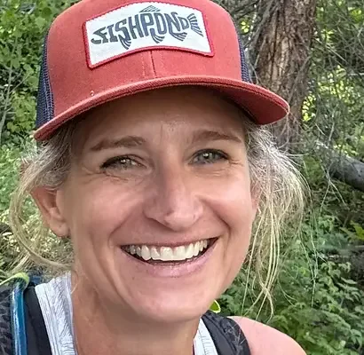 Smiling woman in outdoor setting wearing a red and navy trucker hat with a logo, green foliage in background.