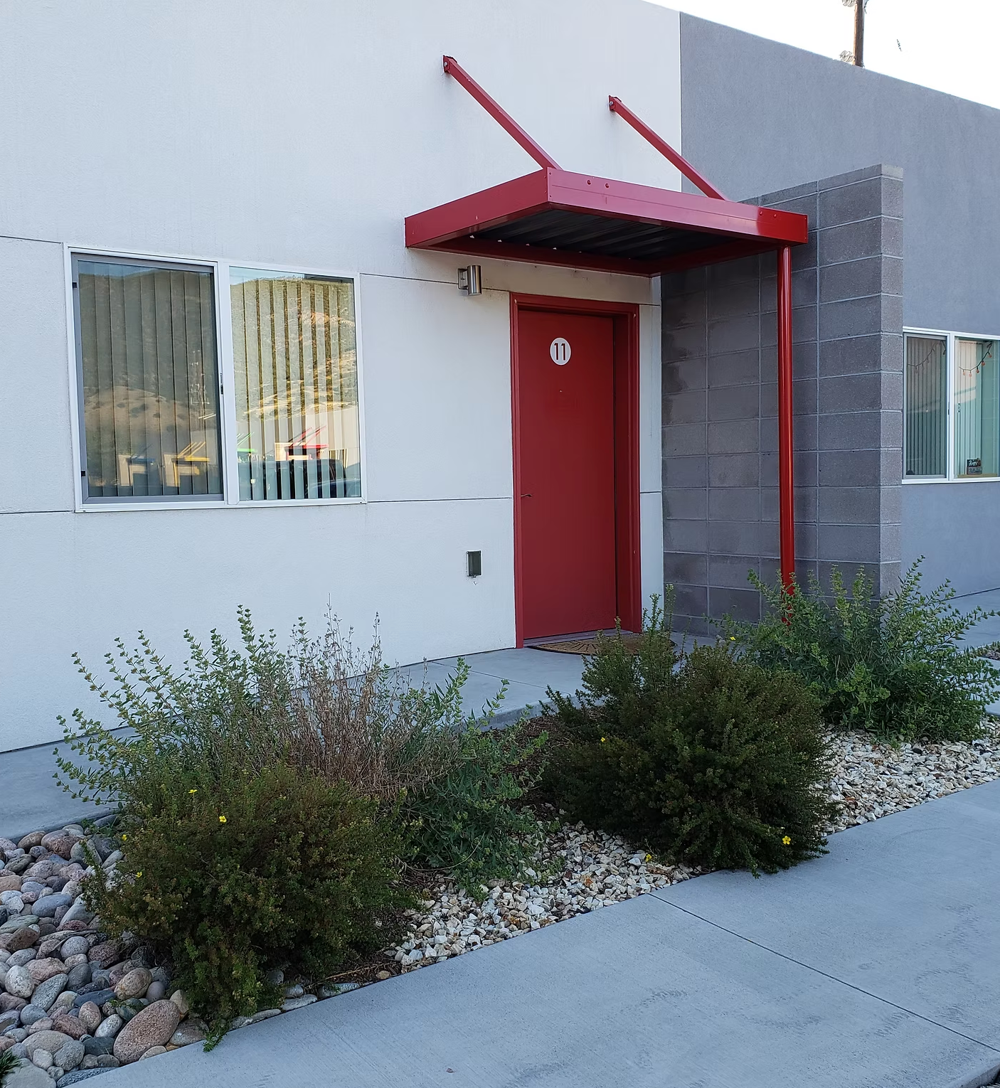 Exterior view of a modern building with a white and gray facade, a red door labeled with the number 11, and a red awning above the door. There are large windows with blinds and small shrubs in a landscaped area with rocks in front.