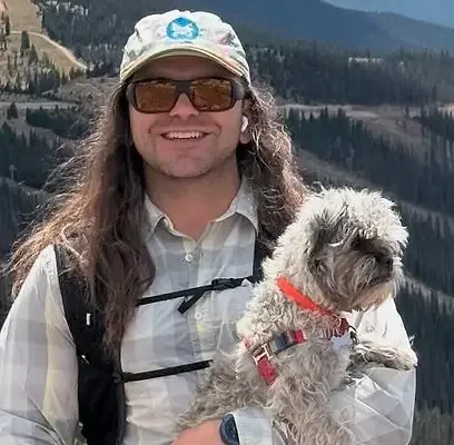 A man with long hair and glasses holding a small dog outdoors in a mountainous area.