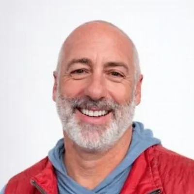 A smiling, bald man with a white beard, wearing a red jacket and a blue shirt, against a plain white background.