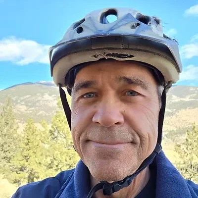 A man wearing a bike helmet outdoors with a mountainous landscape and blue sky in the background.