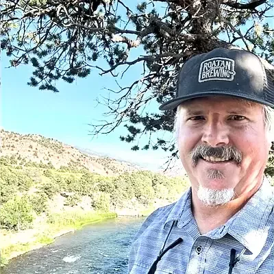 A man with a beard and mustache wearing a black hat that says 'Raftan Brewing' smiling outdoors with a river, trees, and hills in the background.