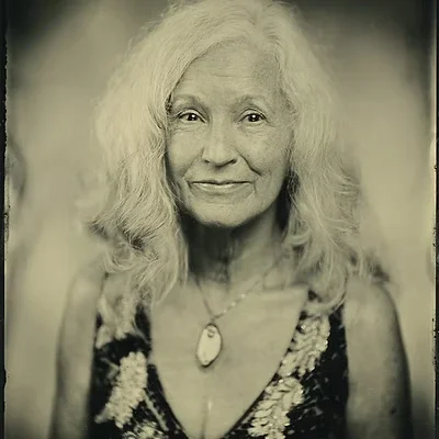 A portrait of an elderly woman with long, wavy gray hair, wearing a sleeveless dress and a necklace with a large pendant.
