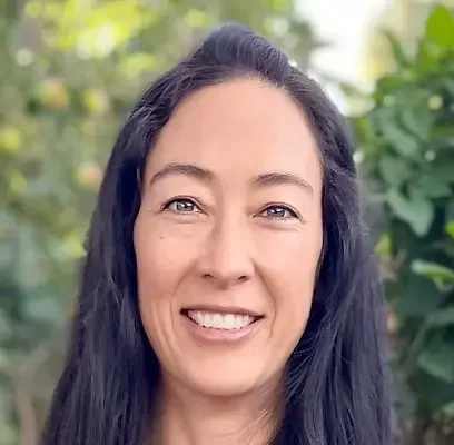Close-up of a smiling woman with long dark hair outdoors, surrounded by green foliage.