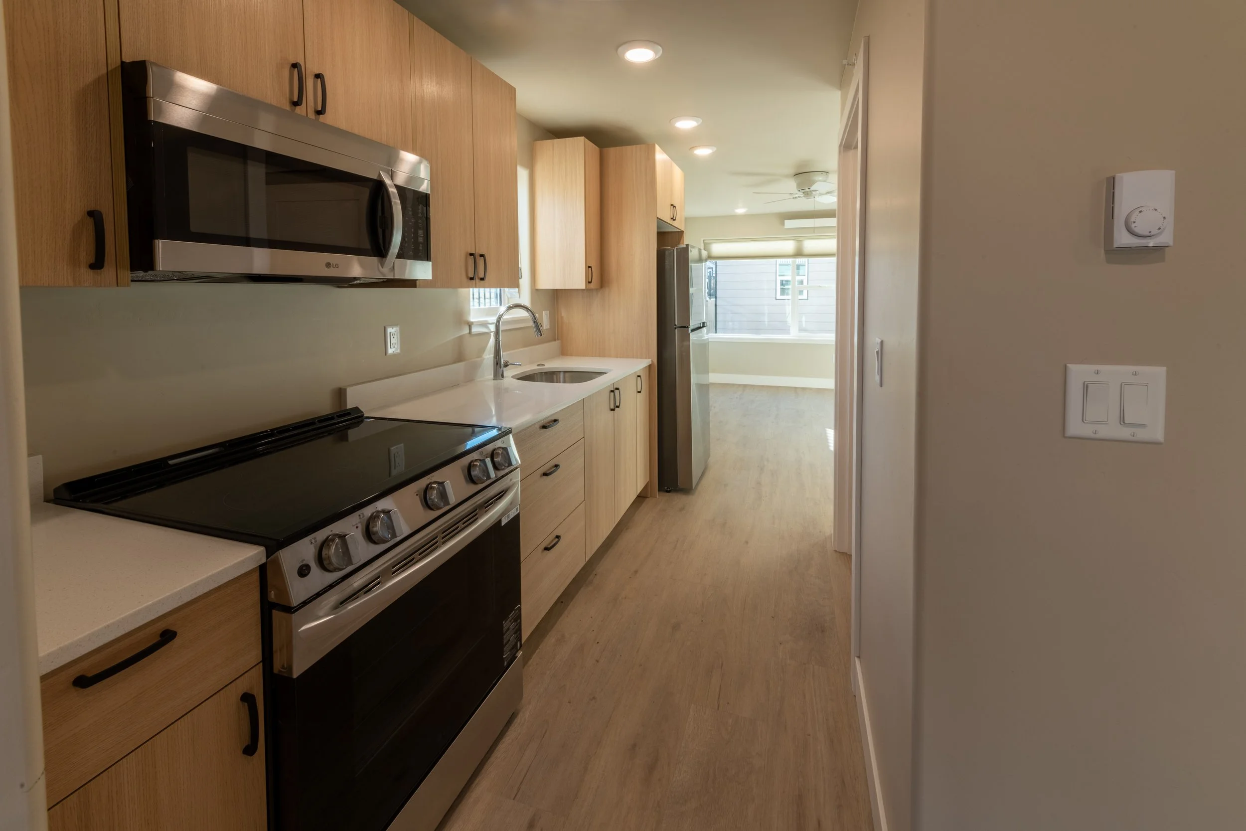 Kitchen with wooden cabinets, stainless steel microwave, stove, refrigerator, and a sink under a window, with hardwood flooring and recessed lighting.