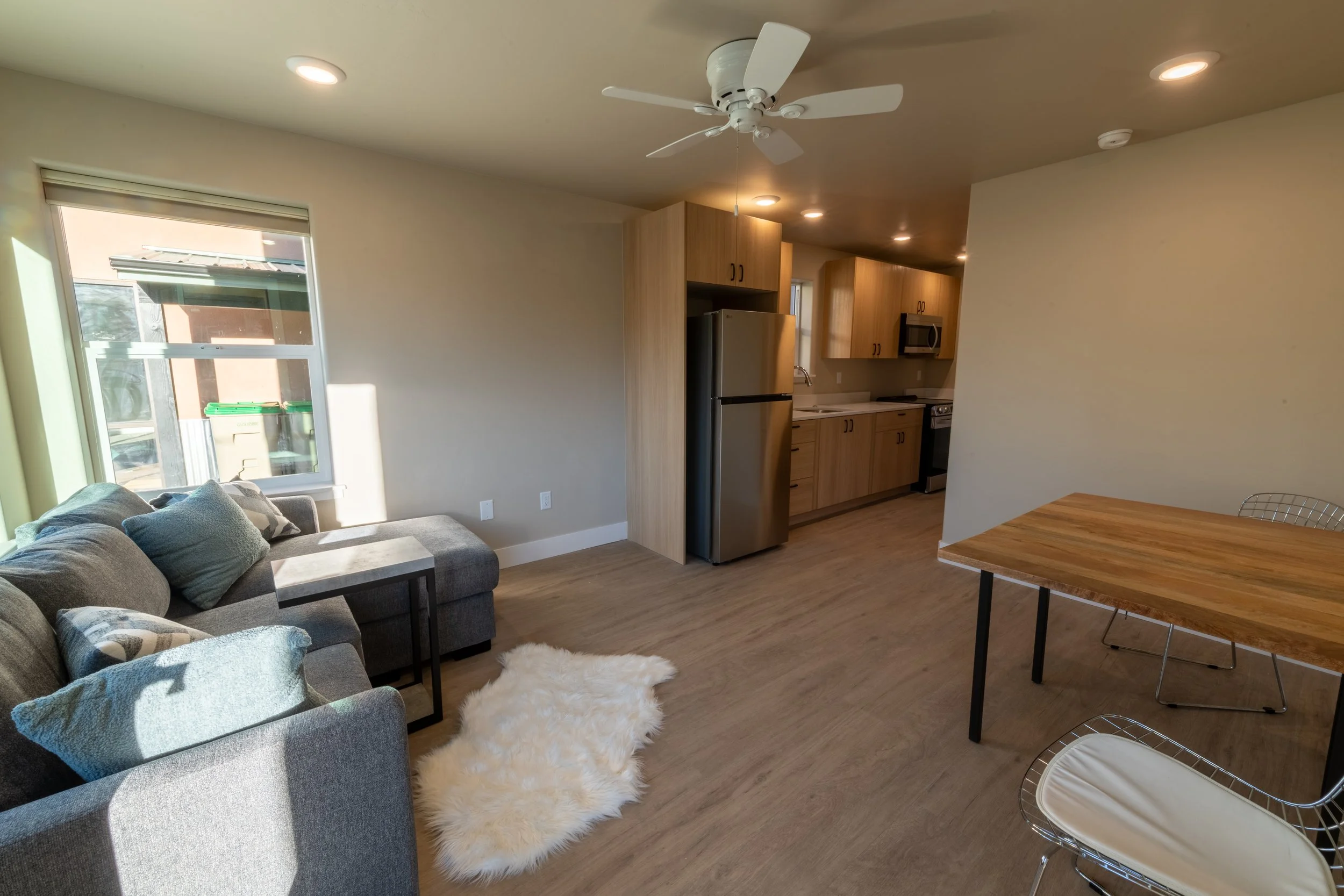 Living room area with a gray sofa adorned with patterned pillows, a wooden dining table with metal chairs, a plush white shaggy rug, and a kitchen with wooden cabinets and stainless steel appliances including a refrigerator, microwave, and stove. A ceiling fan is installed on the ceiling, and large windows let in natural light.