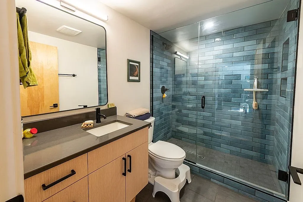 Modern bathroom with a wooden vanity, gray countertop, large mirror, glass shower with blue tile walls, and a toilet with a small step stool.
