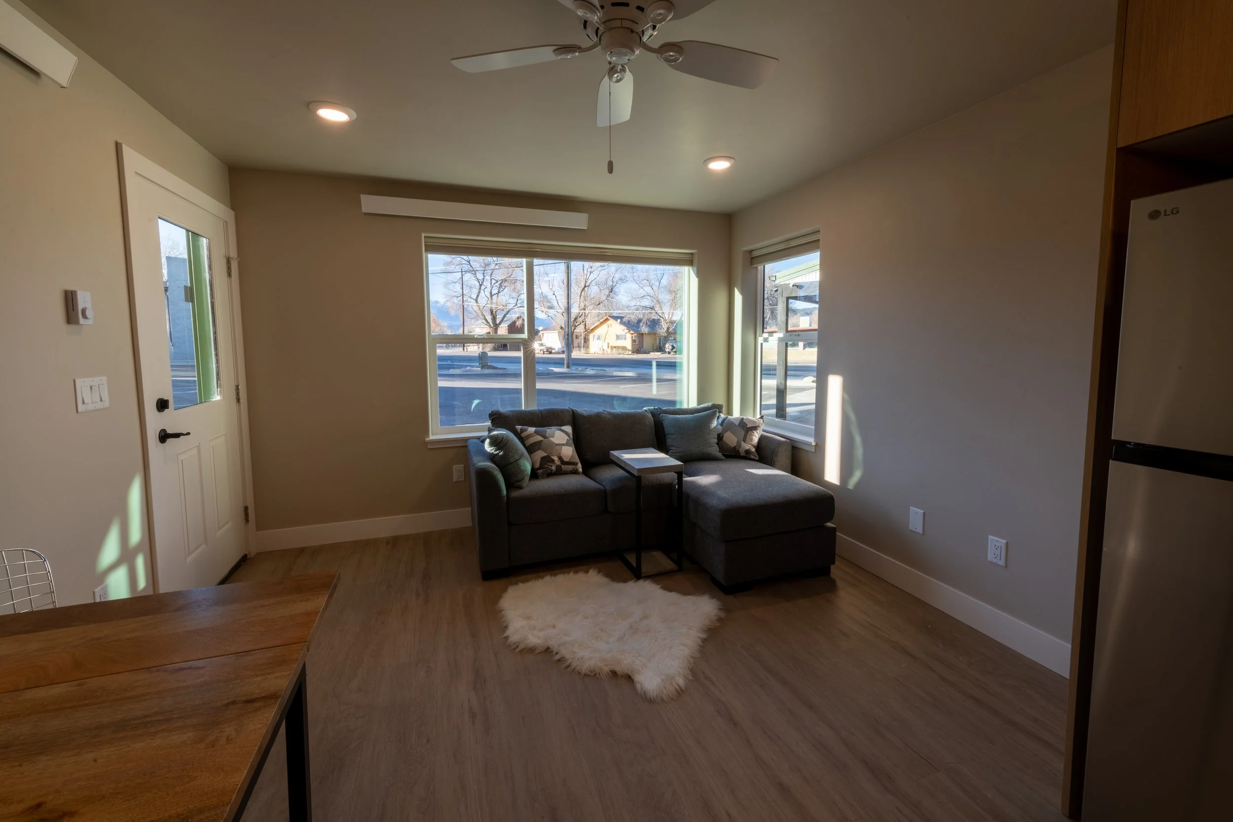 Living room with a gray sofa, large windows, wooden floor, ceiling fan, and white walls.