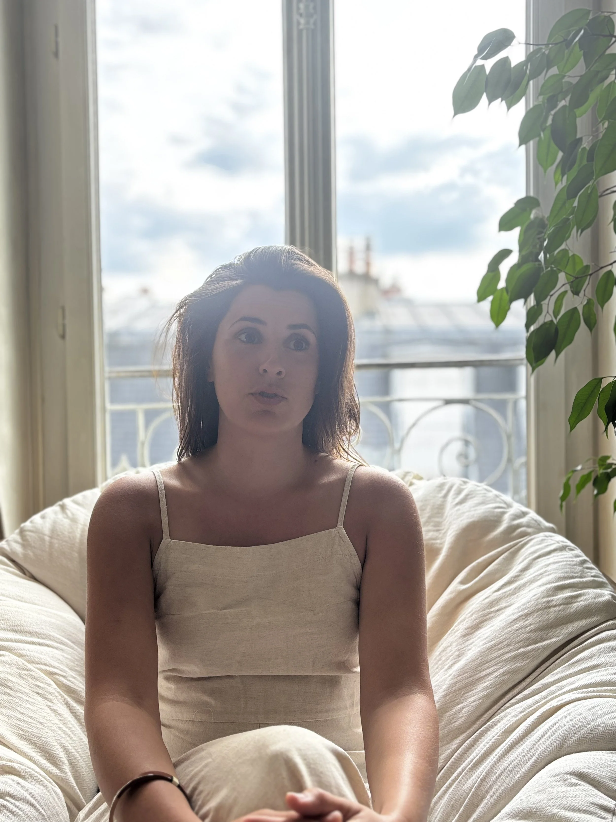 A woman with shoulder-length brown hair sitting on a bed in front of a window with a view of rooftops and a cloudy sky, with a large green plant to her right.