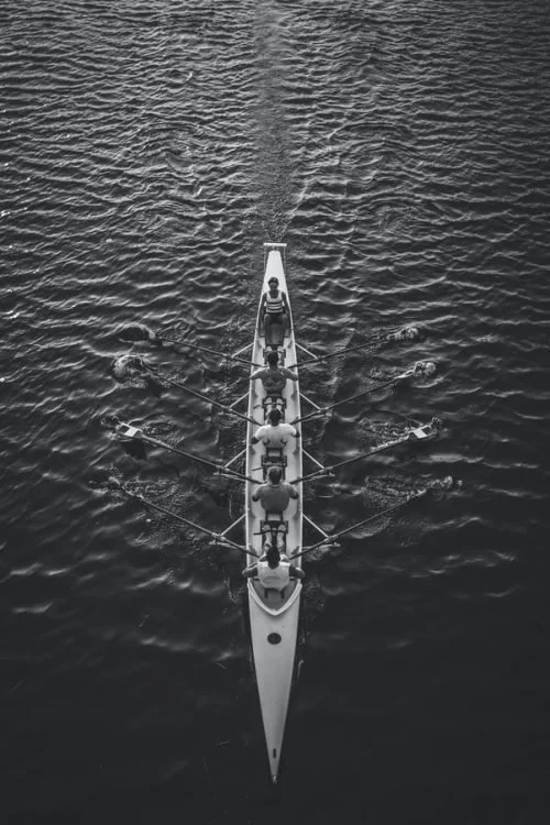 A black and white aerial view of a rowing team practicing on open water, with six rowers in a long boat, synchronized rowing, creating ripples in the water.