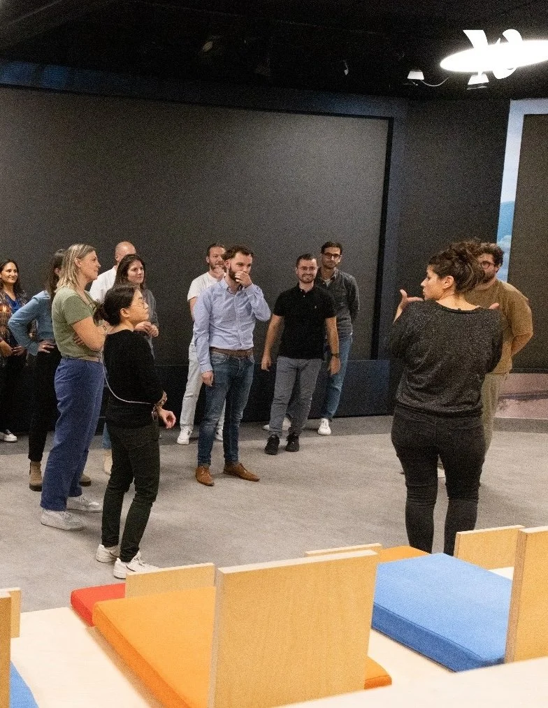 A group of people standing in a semi-circle in a modern room, listening to a woman who appears to be leading a discussion or presentation.