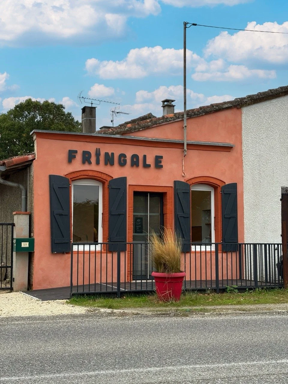 Façade de bâtiment rose avec deux fenêtres à volets noirs et une porte centrale, le tout entouré d'une petite barrière noire, un pot rouge avec des plantes en façade, et un panneau indiquant «FRINGALE». Le ciel est bleu avec des nuages blancs.