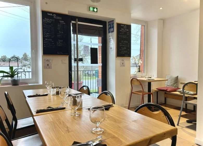 Intérieur d'un restaurant avec une table en bois prête pour des clients, ornée de verres et couverts, et une ambiance lumineuse naturelle
