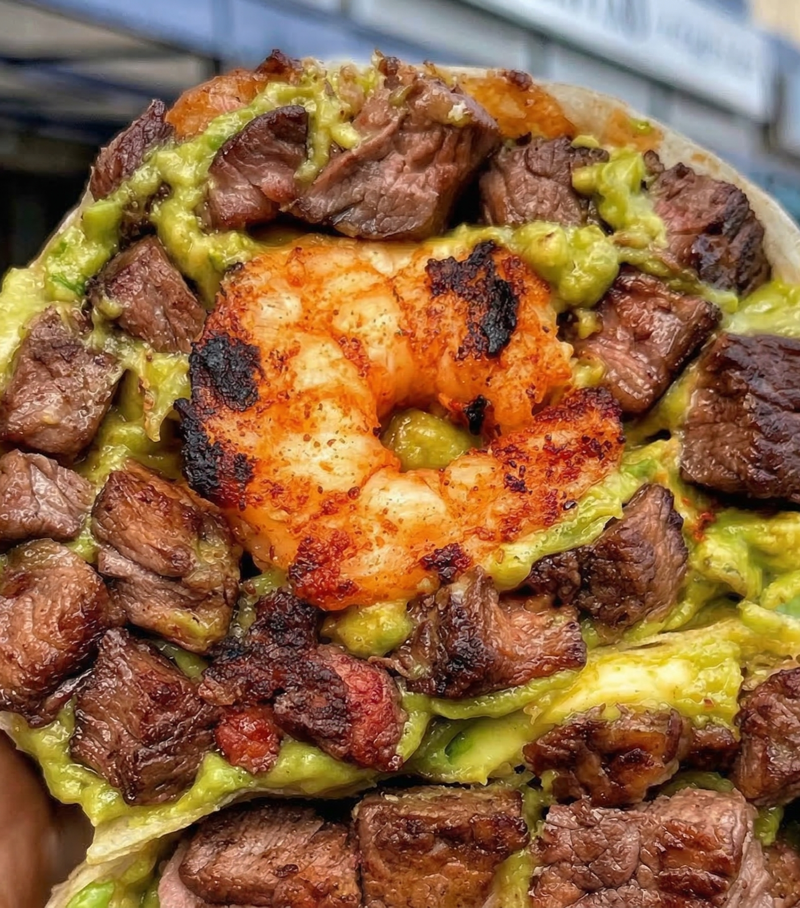 Close-up of a seafood taco with grilled beef pieces, a cooked shrimp, shredded lettuce or cabbage, and guacamole in a soft tortilla.