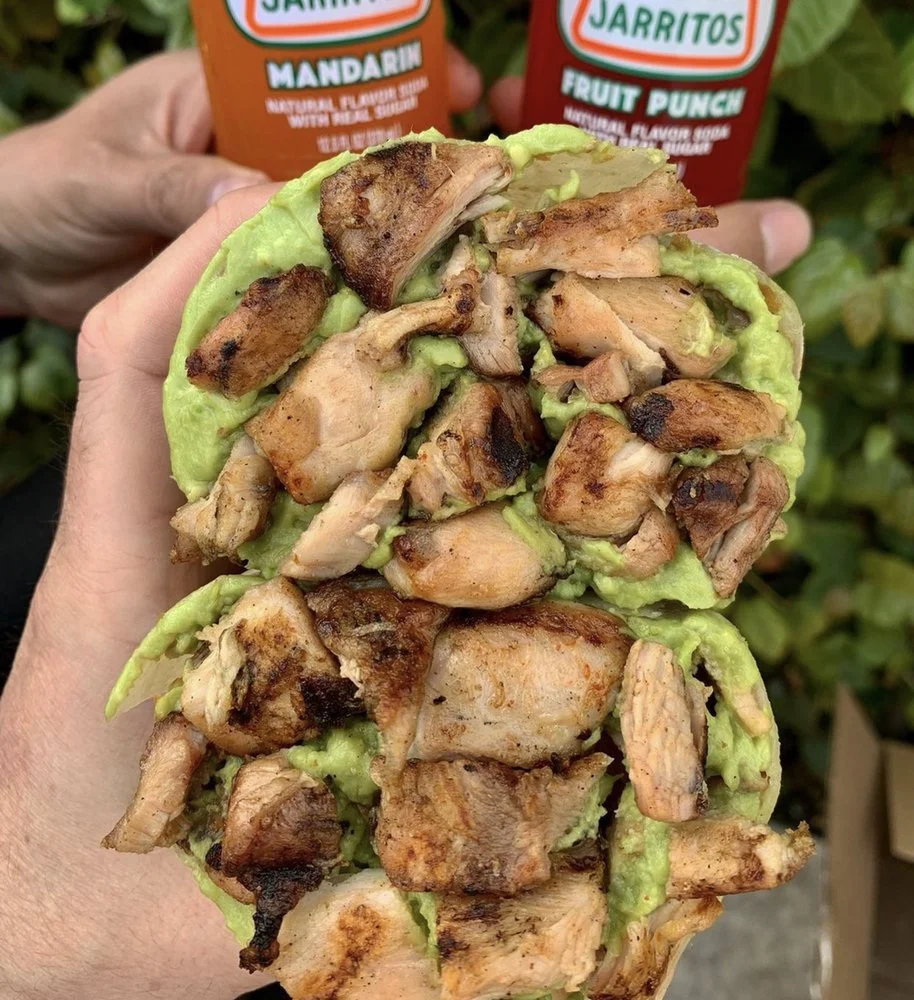 Close-up of a hand holding a taco with grilled chicken pieces on a bed of lettuce. In the background, two bottles of Jarritos soda, one Mandarin and one Fruit Punch, are visible.