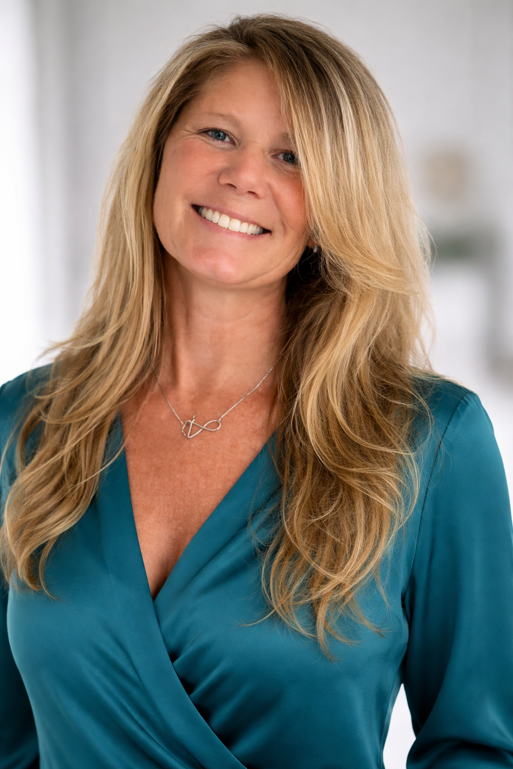A woman with long blonde hair smiling, wearing a teal blouse and a silver necklace with a heart and infinity symbol design.