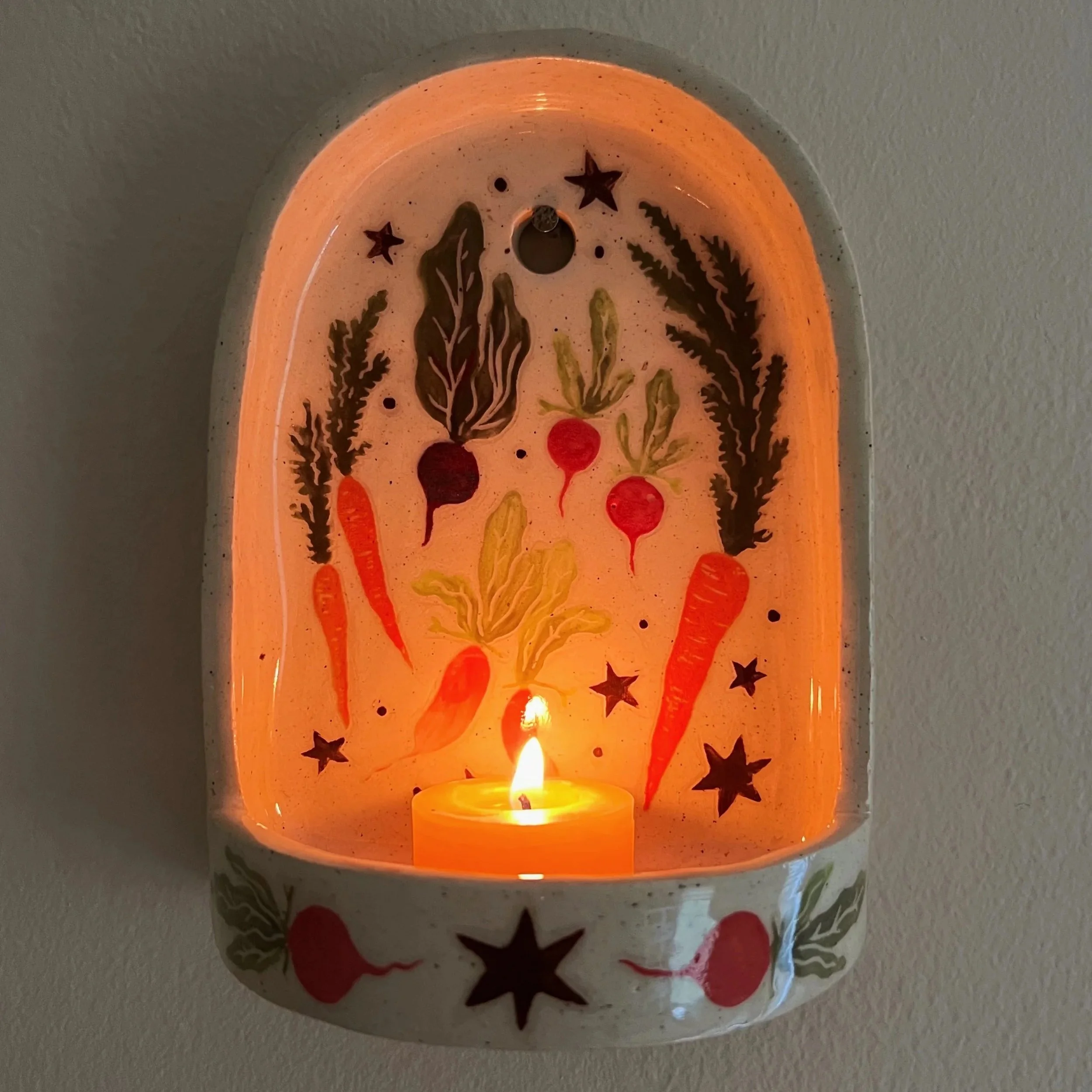 A wall-mounted candle sconce with a glowing candle inside, featuring painted vegetables and stars.