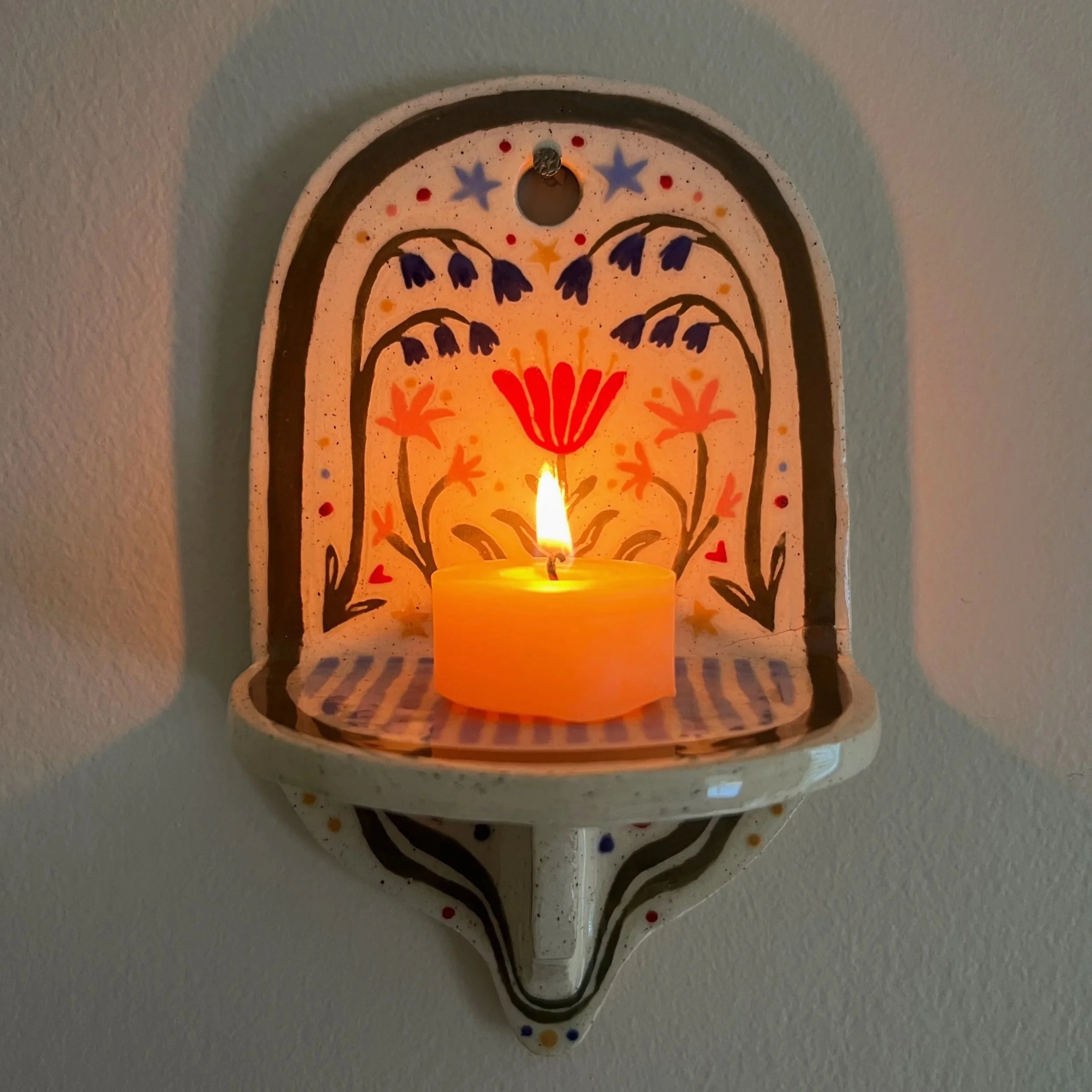 Decorative wall-mounted candle sconce with painted floral and star designs, illuminated by a lit beeswax candle.