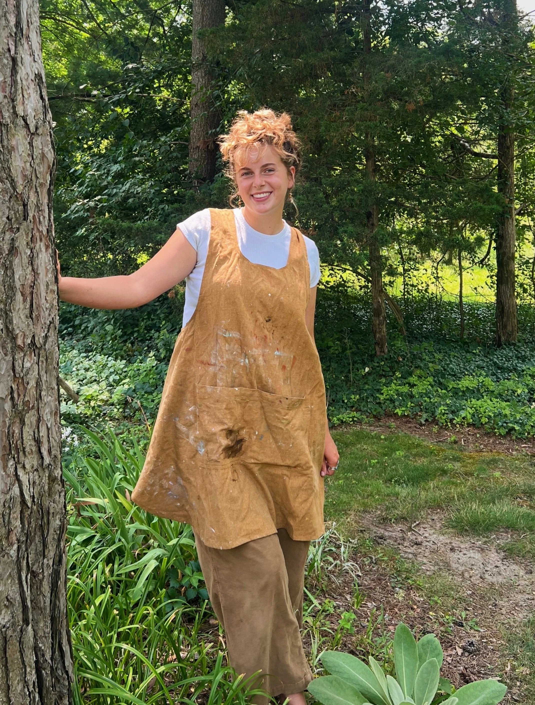 Claire, a woman with curly blonde hair wearing a yellow apron over a white t-shirt, is standing outdoors in a lush, green garden next to a tree. Her apron has paint splatters on it.
