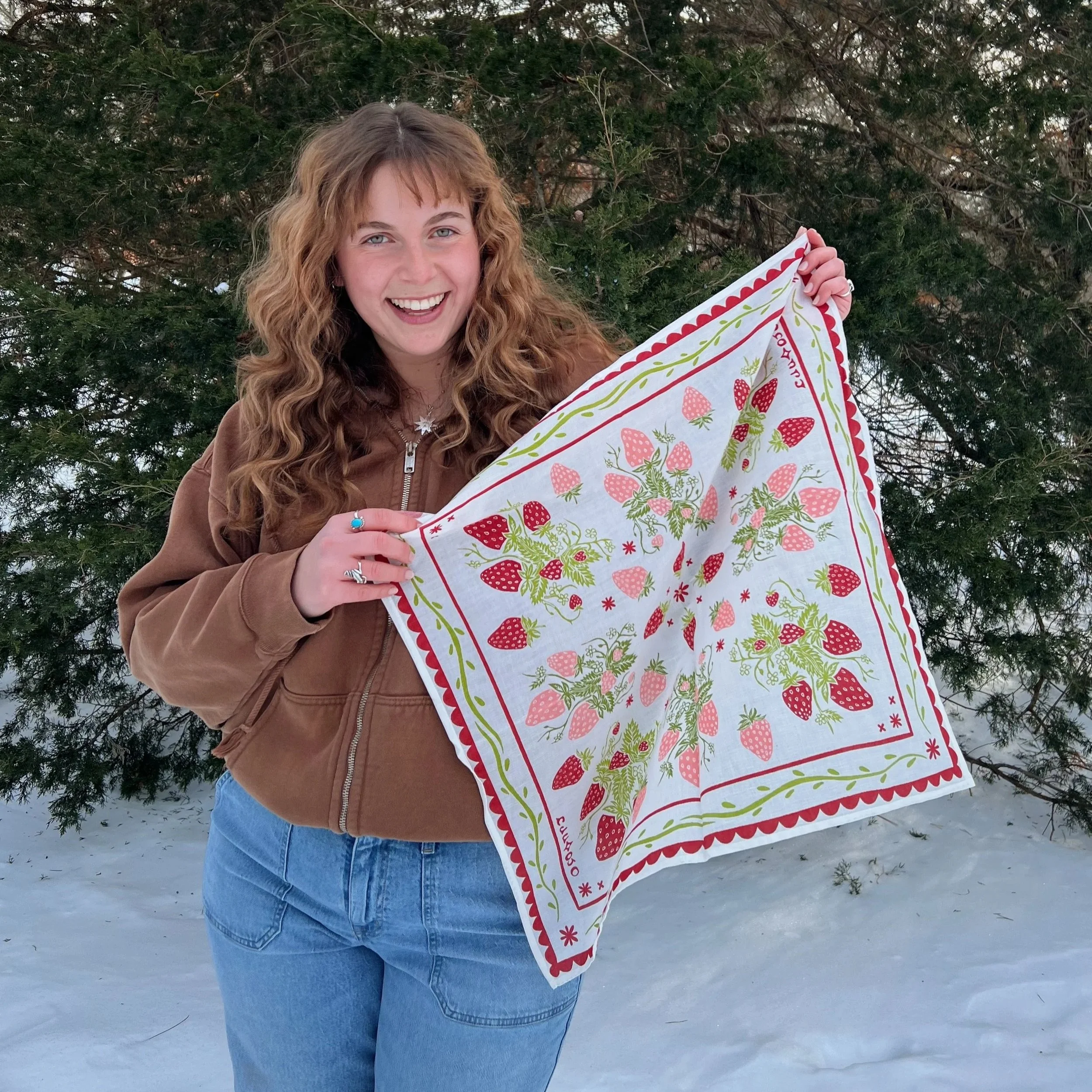 A woman with curly blonde hair smiling outdoors in winter, holding a colorful strawberry-patterned bandana, with snow on the ground and evergreen trees in the background.