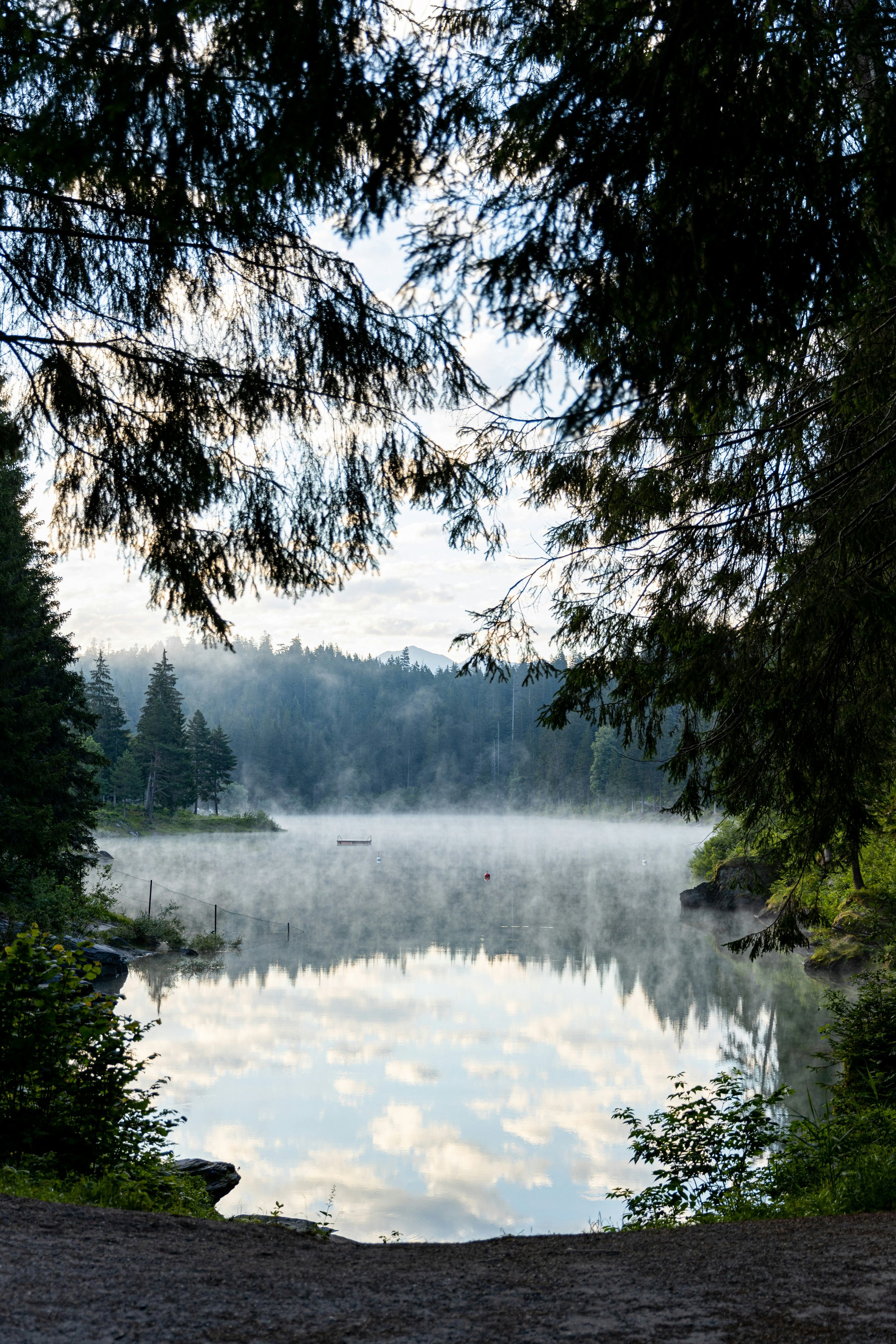 A serene lake surrounded by dense trees, with a small boat on the water and mountains in the background. Mist floats above the water's surface.