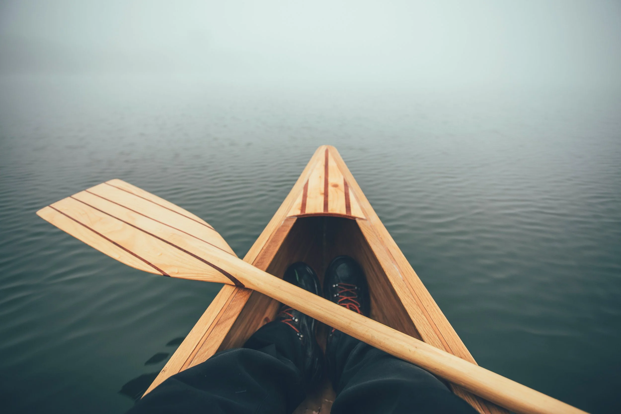 Wooden kayak on calm water with person's legs and shoes visible inside the kayak.