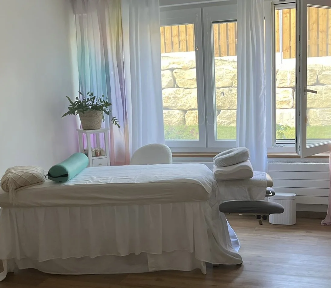 A massage therapy room with a massage table draped in white linens, a lumbar bolster, and folded towels. There's a window with sheer white curtains letting in natural light, overlooking a stone wall and yard outside. A potted plant and a small white shelf are in the corner.