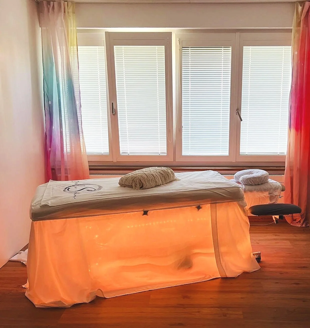 A massage table with a white sheet and cylindrical pillow, covered by a sheer orange fabric that glows from beneath, in front of a large window with closed white blinds and colorful curtains, on a wooden floor. Seelesein . Praxis in Ins Bern Seeland