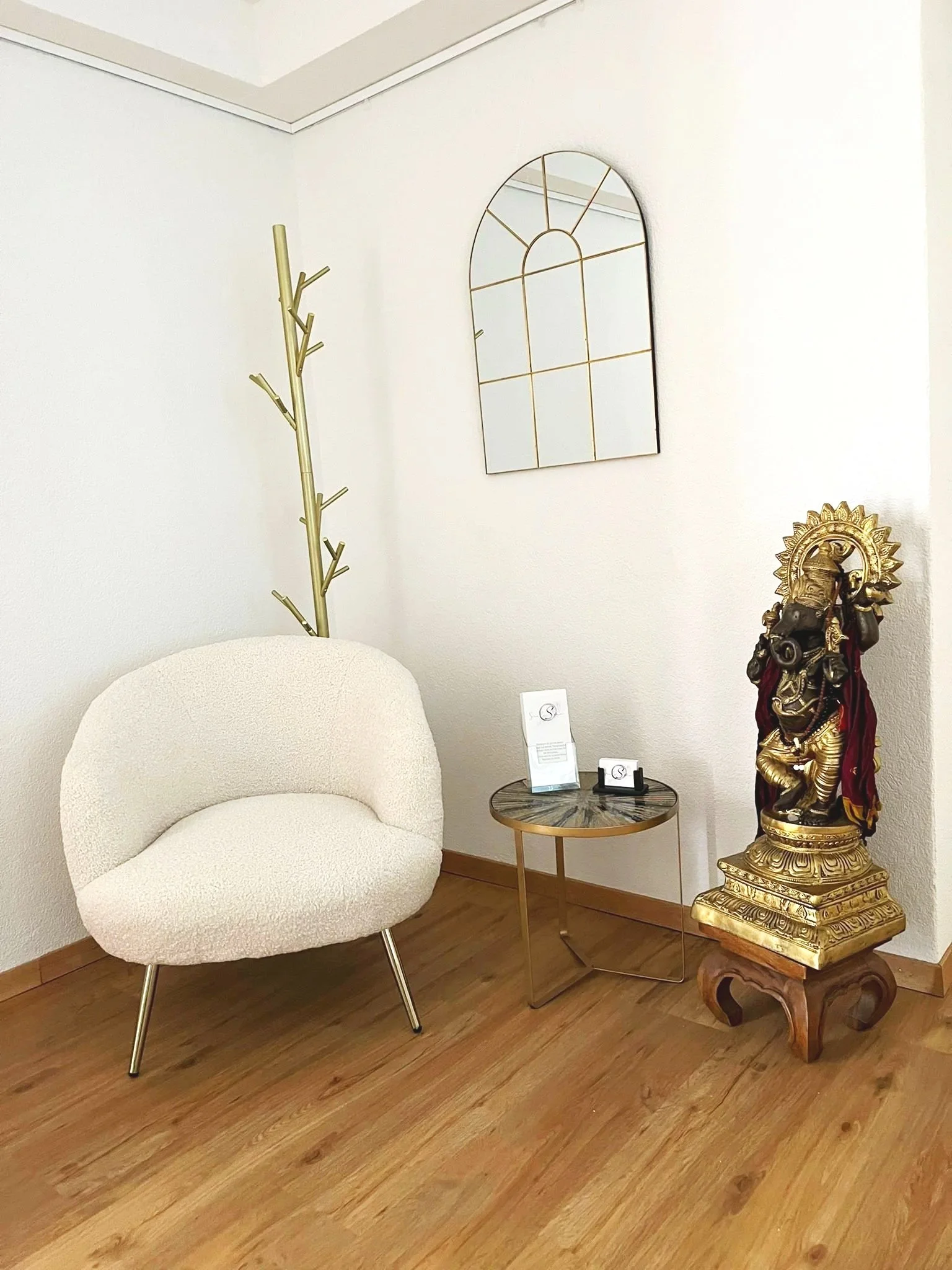 A cozy corner with a modern cream-colored armchair, a small round marble-topped side table holding a brochure and business card, a tall gold coat rack shaped like a tree, a decorative gold and black statue of Ganesha, a white wall with a gold-framed mirror, and wooden flooring.