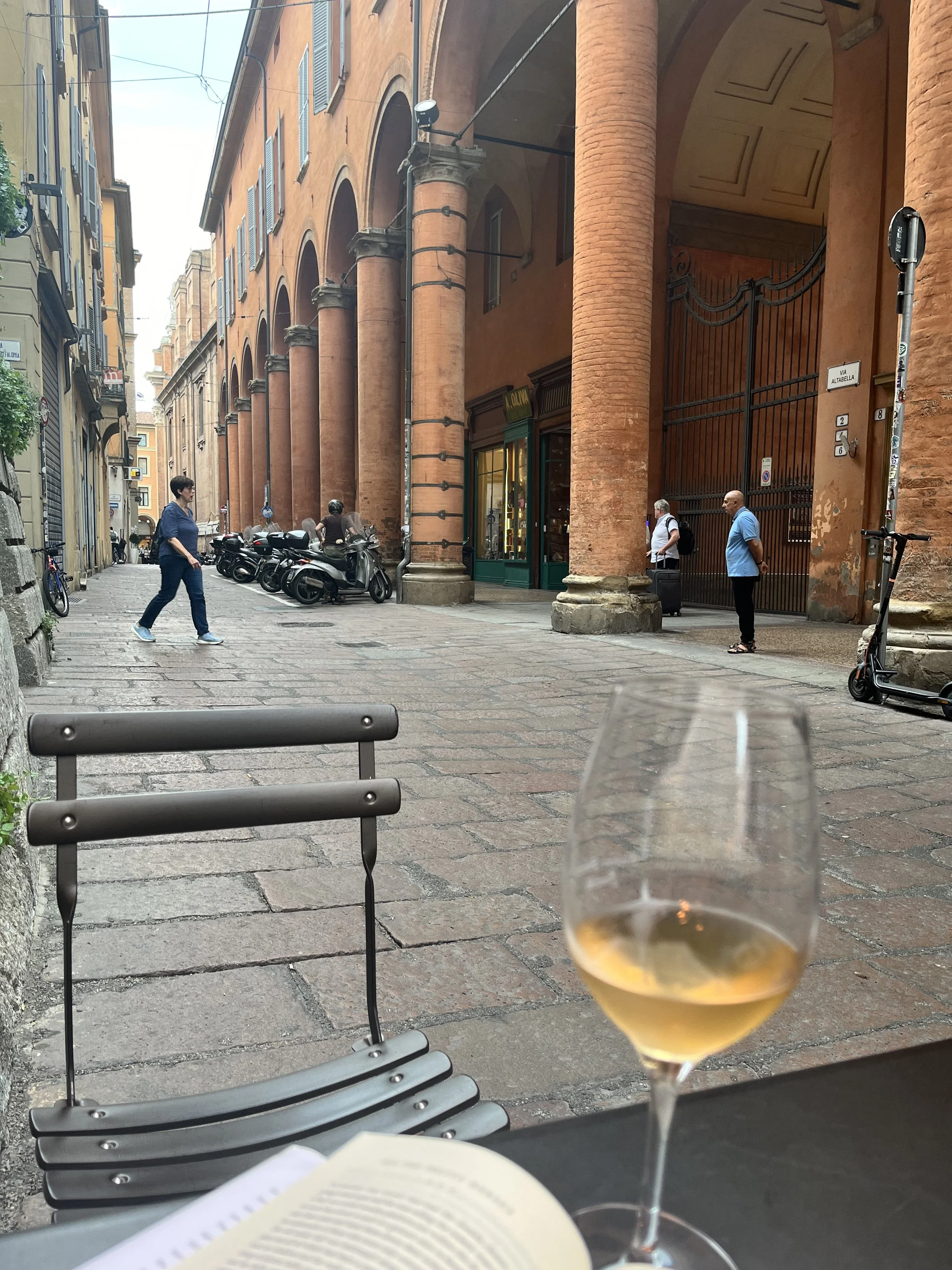 View from a cafe table showing a street scene with pedestrians, motorcycles, and an orange-colored historic building with large columns and arched windows, on a sunny day.
