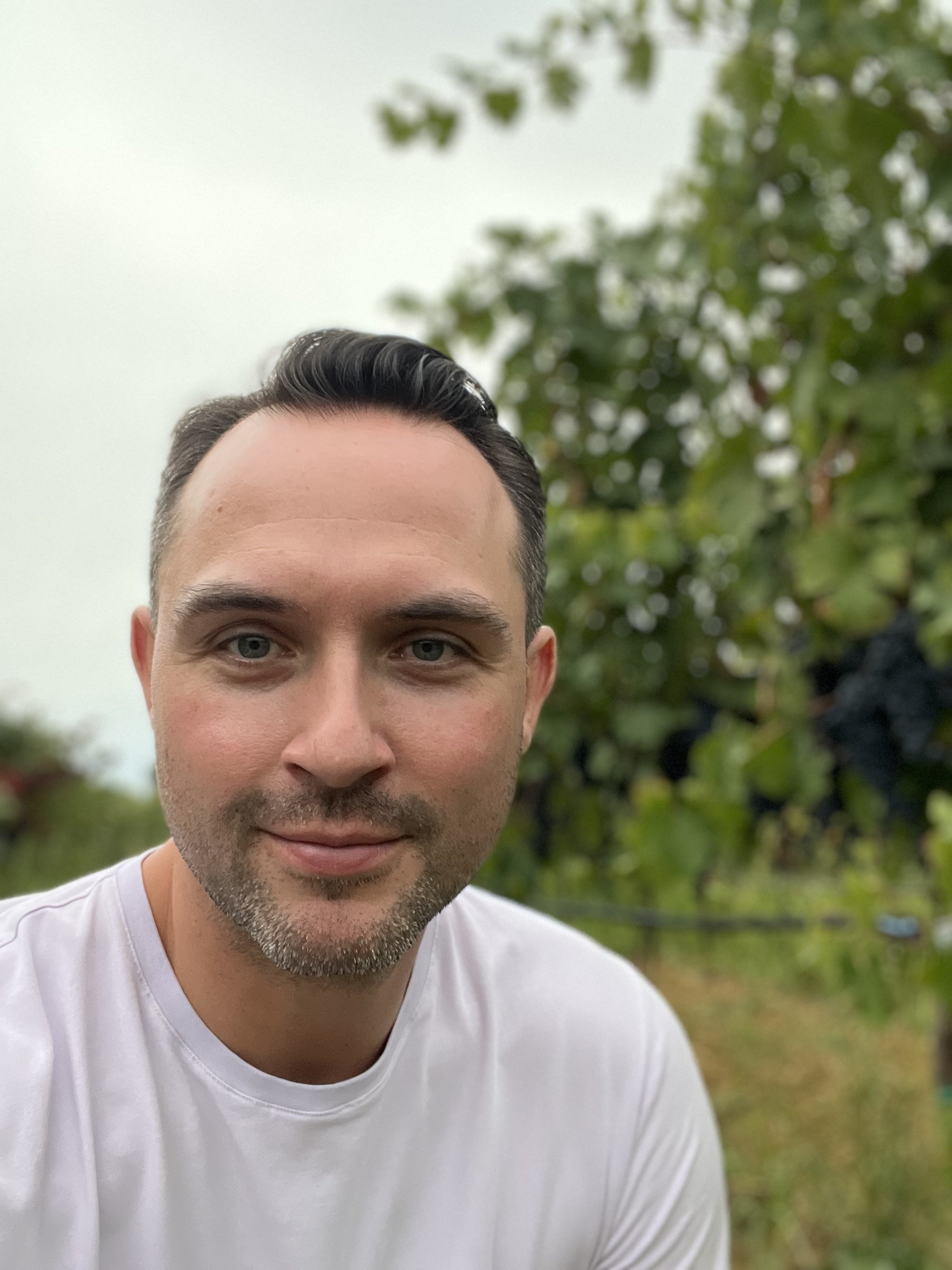 A man with short dark hair and a light beard taking a selfie outdoors, with trees and green foliage in the background.