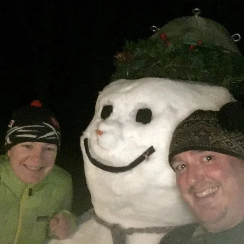 Two men dressed in winter clothing and knit hats taking a selfie with a snowperson that has a smiley face and a Christmas tree in the background.
