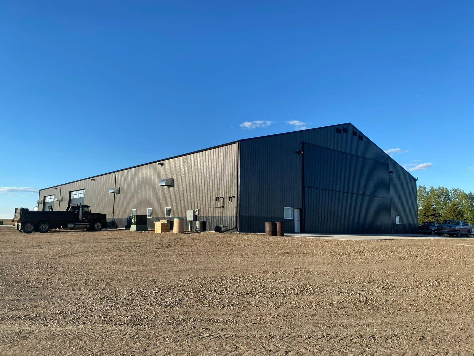A large gray industrial warehouse building with a sloped roof and multiple garage doors, situated on a gravel lot under a clear blue sky with a few clouds, with some parked vehicles and trees in the background.