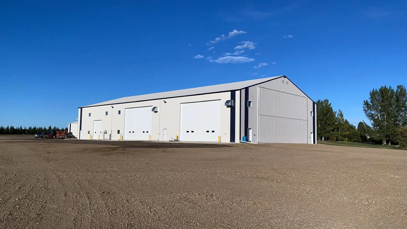 A large white metal building with multiple garage doors sits in a rural area with a clear blue sky and some trees in the background. The ground around the building is bare and unpaved.
