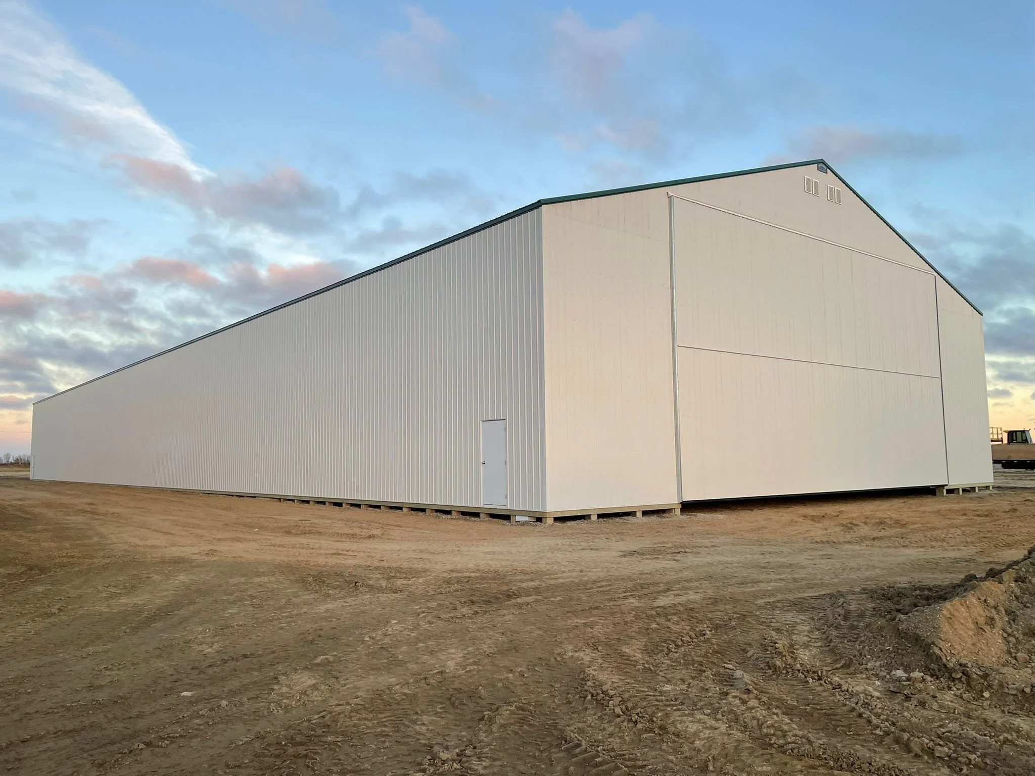 A large white metal storage barn with a sloped roof, situated on a dirt lot during sunset or sunrise.