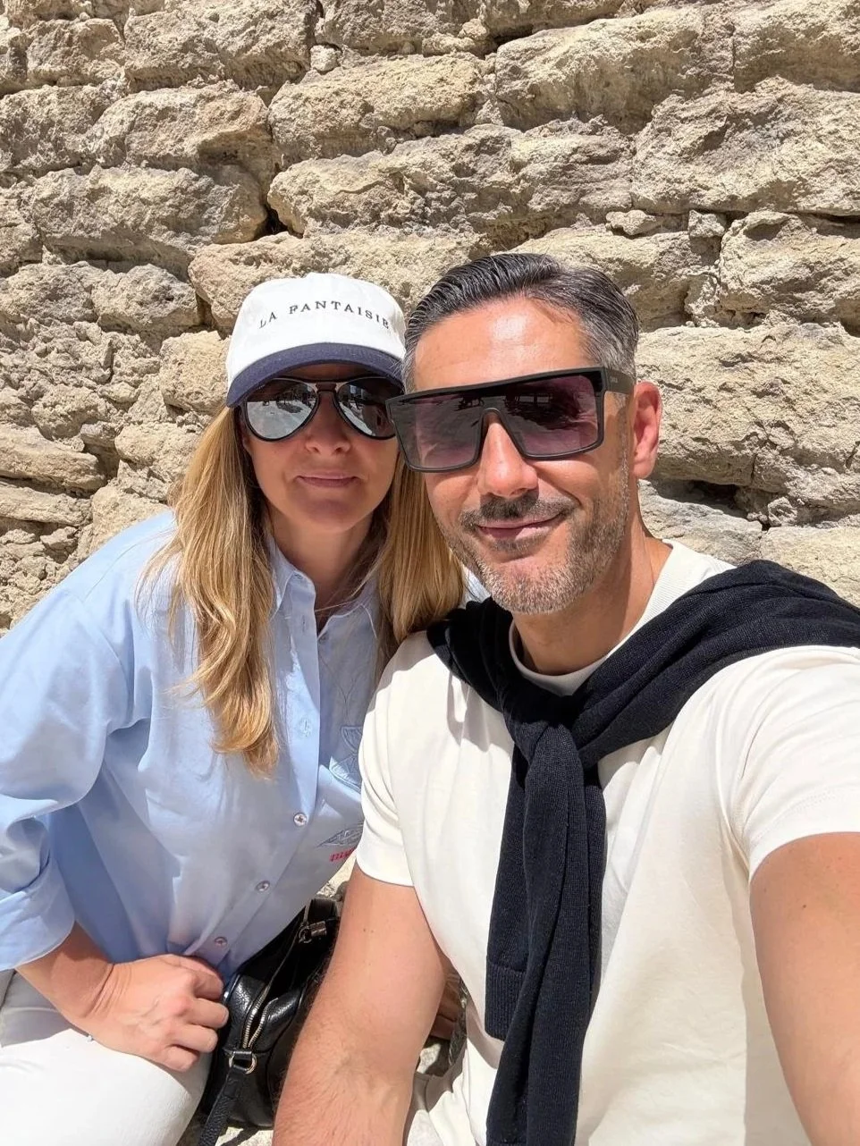 A man and woman taking a selfie outdoors against a rocky wall, both wearing sunglasses and casual clothing, with the woman wearing a white cap and the man with a black sweater draped over his shoulders.