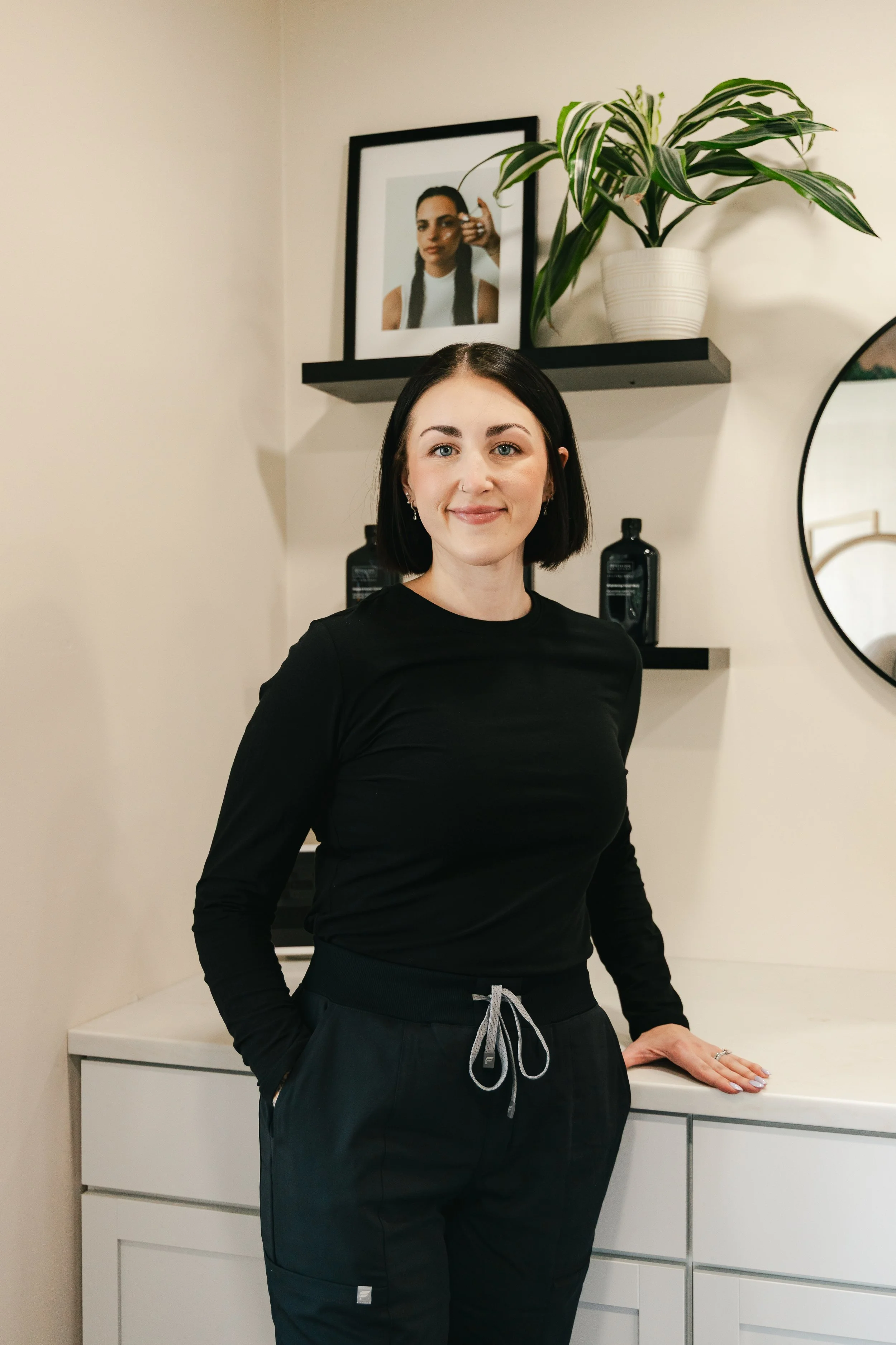 A woman with black hair and pierced ears wearing a black long sleeve shirt and sweatpants, standing in a room with a white dresser and black shelves. There is a framed photo of a woman taking a selfie, and a large potted green plant on one of the shelves.