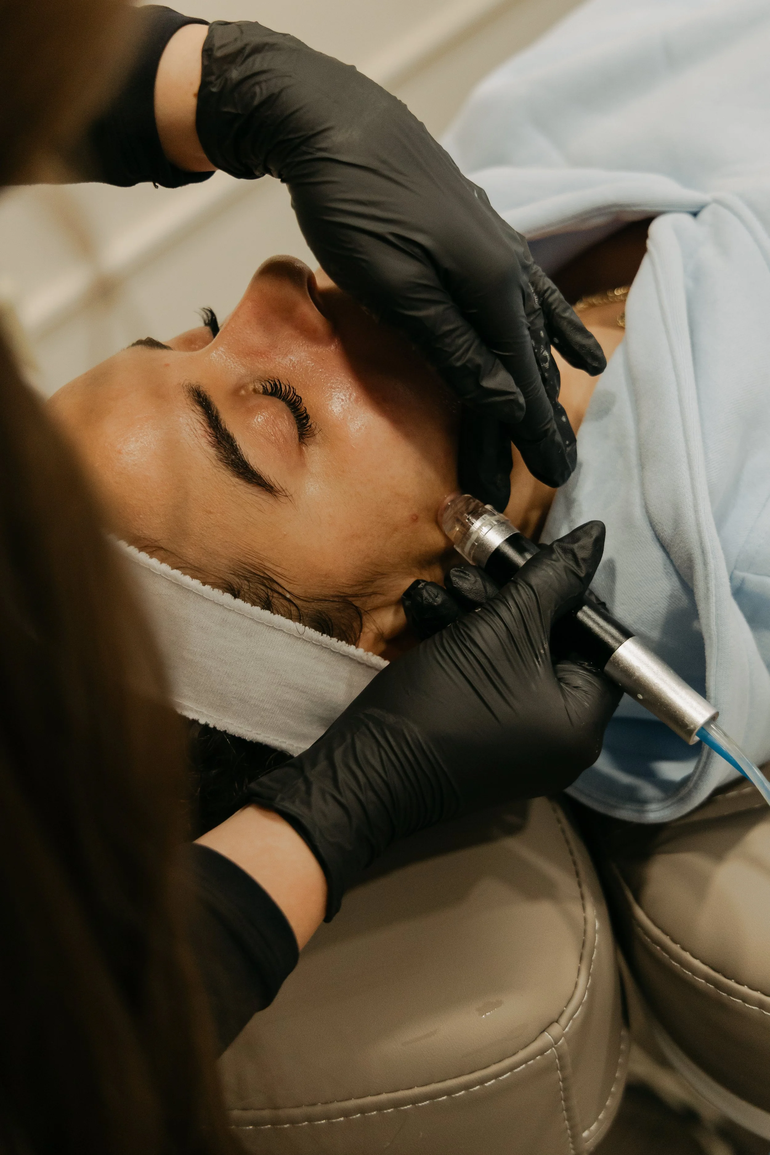 A person receiving a facial treatment with a microdermabrasion device. The person is lying down with their eyes closed, and a skincare professional wearing black gloves is using the device on their face. The person is wearing a white headband and a white garment.