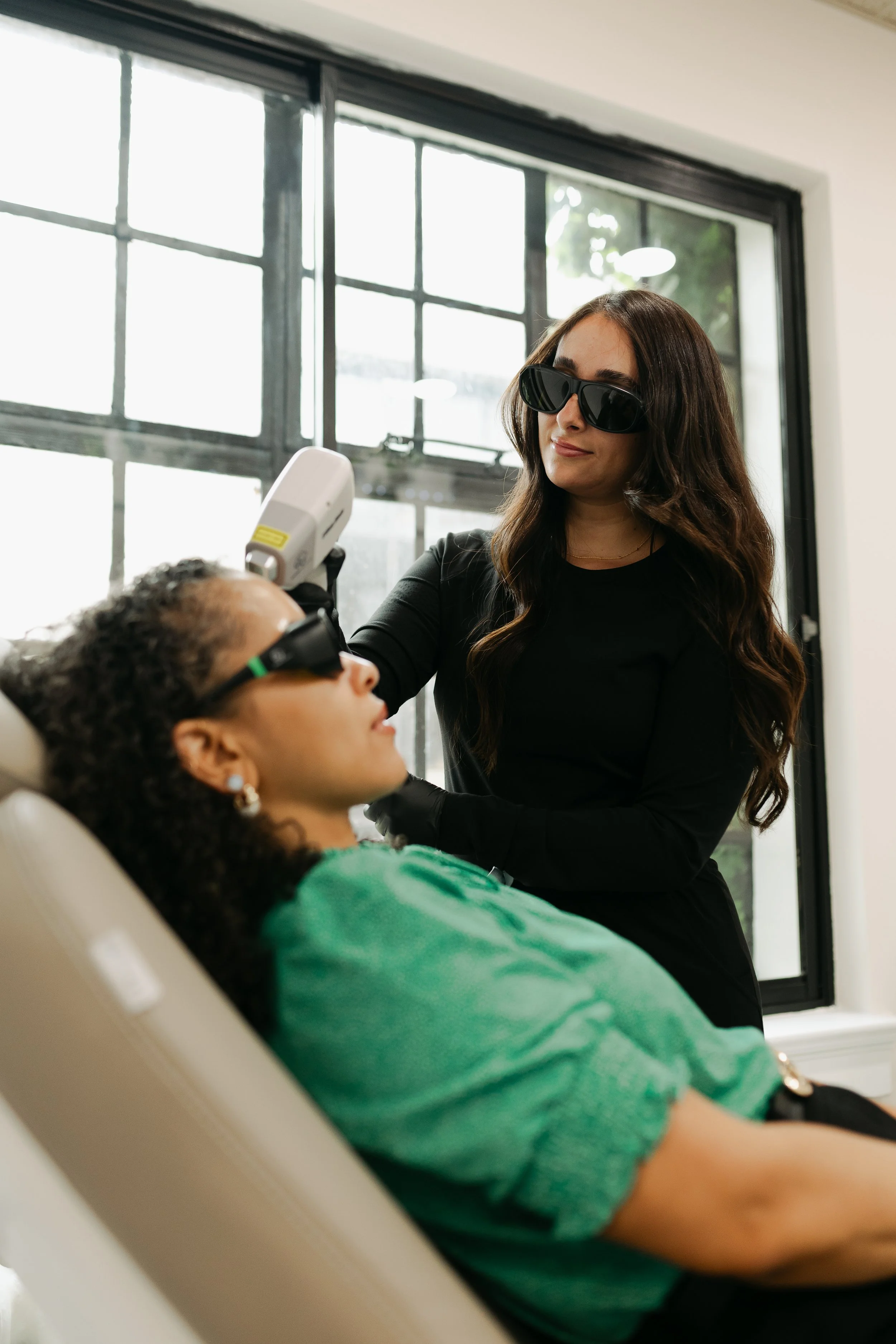 A woman with long dark hair wearing sunglasses stands next to a woman lying back in a dental chair, getting dental treatment, in a room with large windows.