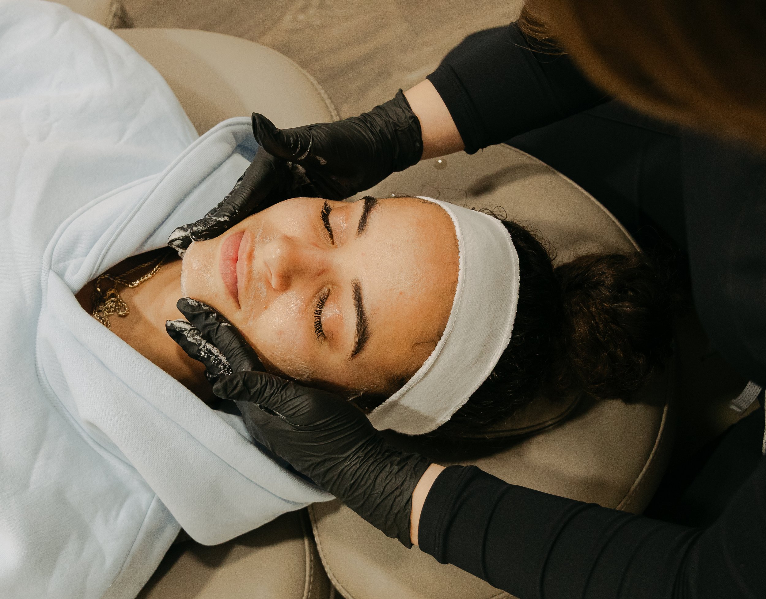 A woman receiving a facial treatment at a spa or beauty clinic, lying on a treatment bed with a white headband and a skincare product applied to her face, while a professional in black gloves gently massages or applies skincare to her face.