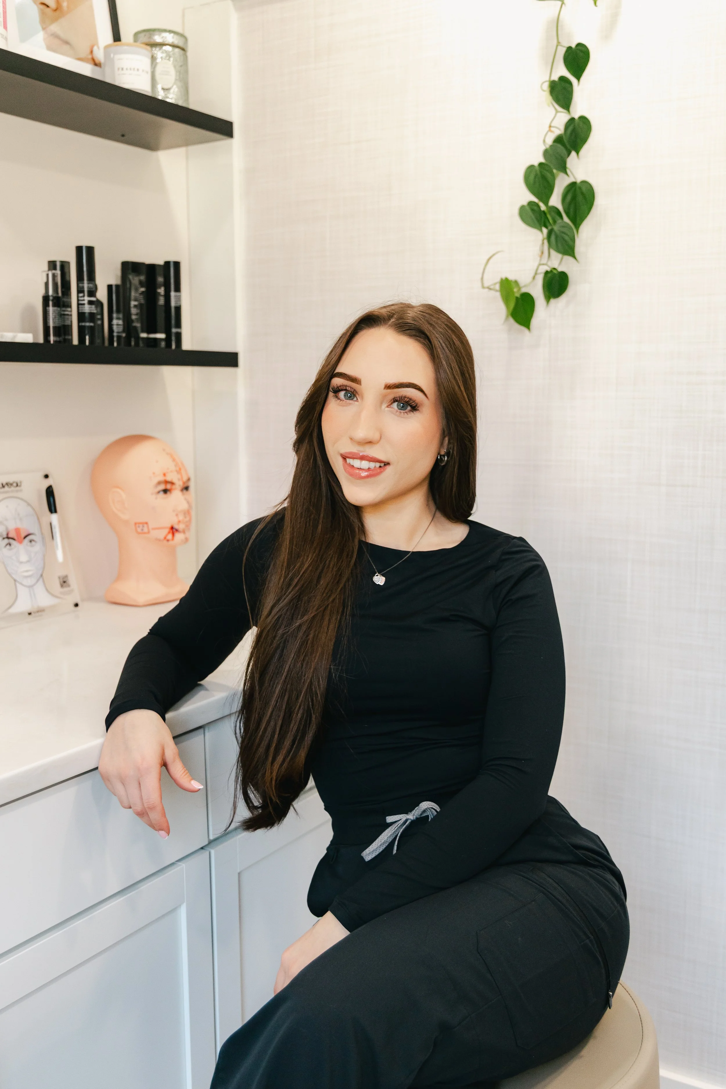 A woman with long brown hair and blue eyes sitting on a beige stool, wearing a black long-sleeve shirt and black pants, smiling at the camera in a medical or therapy office with a mannequin head and facial diagram on a shelf behind her, and a green plant hanging on the wall.