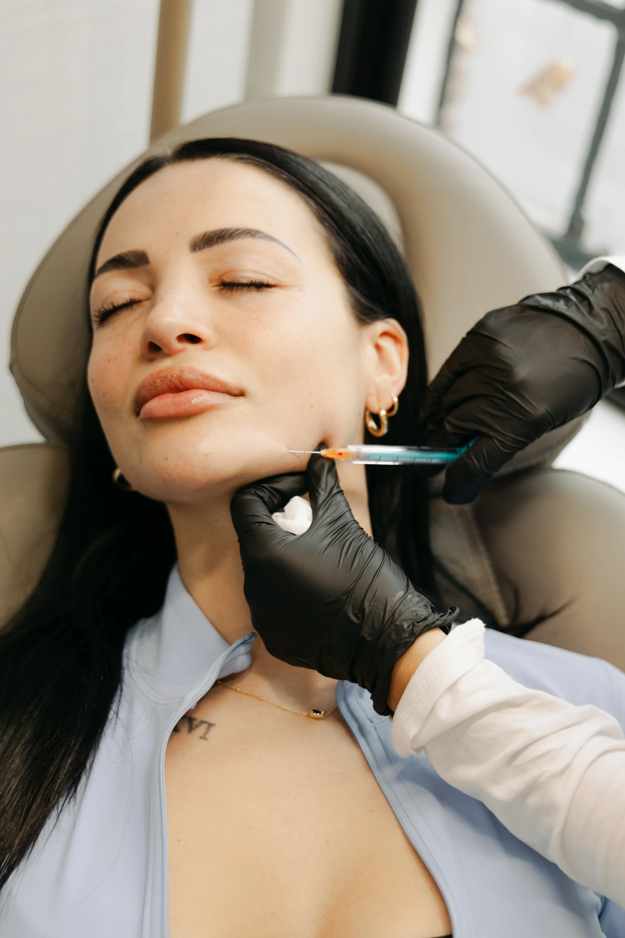 A woman with closed eyes receiving a cosmetic injection in her face from a medical professional wearing black gloves.
