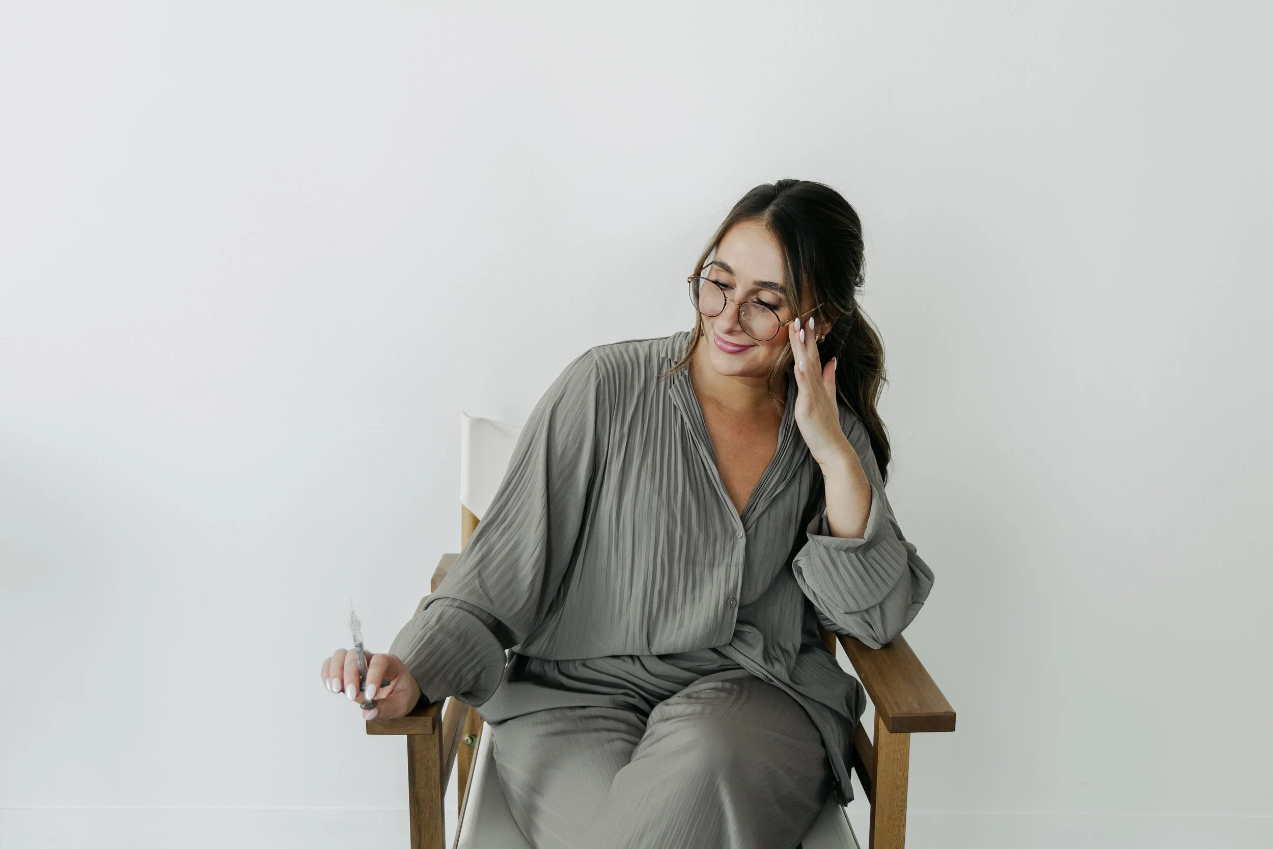 A woman with glasses and a nose ring sitting on a wooden chair against a plain white wall, holding a pen, with her hand resting on her face, smiling with her eyes closed.