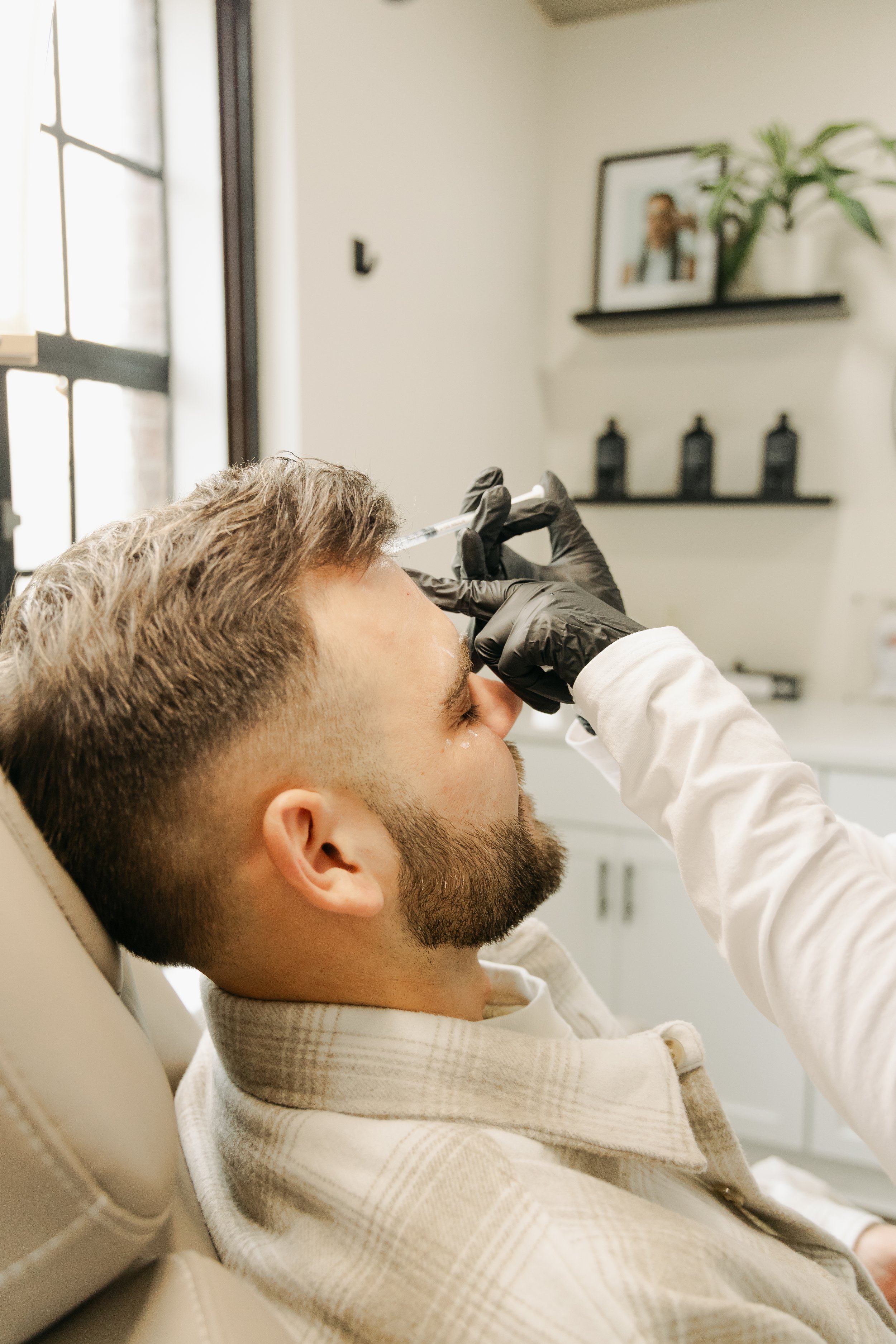 A man with a beard wearing a white shirt is lying in a dental chair, receiving a dental injection from a dental professional wearing black gloves. The background shows a shelf with framed pictures and decorative items in a well-lit dental office.